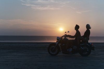 People riding motorcycle on beach against sky during sunset
