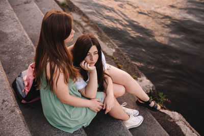 Girlfriends embracing while sitting on steps by river in city