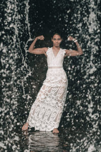 Portrait of woman wearing dress flexing muscles while standing by waterfall