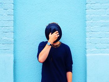 Teenage boy hiding face with hat while standing against blue wall