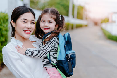 Portrait of happy mother carrying schoolgirl on footpath