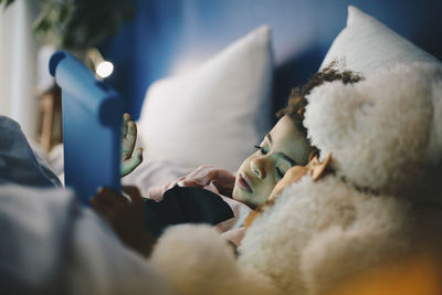 Boy lying with teddy bear while using digital tablet on bed at home