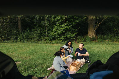 Family having snacks while sitting on grass at park seen through car trunk