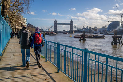 Rear view of people walking on footpath by tower bridge over thames river against sky