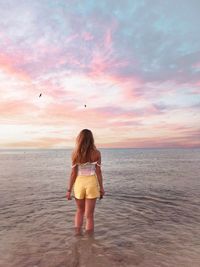 Rear view of woman standing in sea against sky during sunset