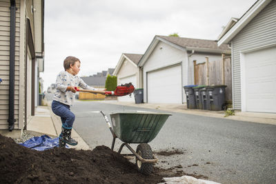 Young boy lifting shovel of dirt into wheelbarrow in back alley