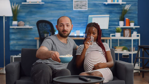 Pregnant couple watching tv while holding drink and popcorn on sofa at home