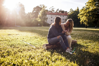 Mother talking to daughter while sitting on log amidst grassy field at park