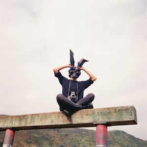 Man wearing mask sitting on retaining wall against sky