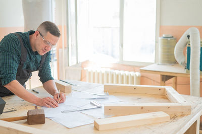 A portrait of a carpenter in goggles and work overalls draws a blueprint for a workshop