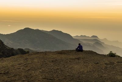 Man sitting on mountain against sky during sunset