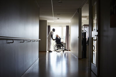 Full length side view of male nurse pushing senior man on wheelchair at hospital corridor