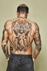 Back view of unrecognizable man with muscular tattooed body in jeans standing against beige background