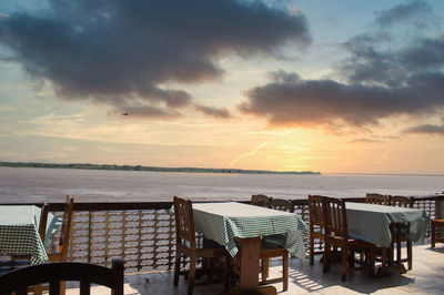 Empty chairs and table by sea against sky during sunset