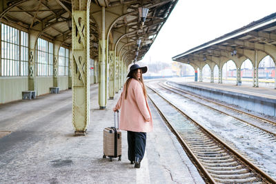 Young woman in coat walks with suitcase along empty platform of railway