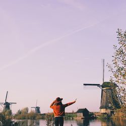 Rear view of woman standing by river with traditional windmill against sky