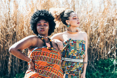 Confident black and mixed race female models in ornamental dresses with authentic african accessories looking at camera against dry grass in sunlit field