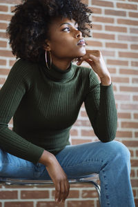 Young woman looking away while sitting on seat against wall
