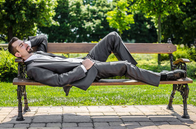 Full length of bridegroom in suit sleeping on bench at park