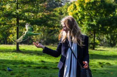 Young woman looking at bird while standing against trees in park