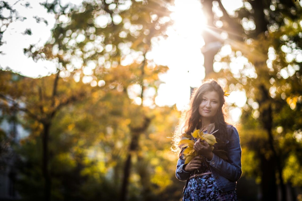 lifestyles, young adult, person, casual clothing, leisure activity, tree, focus on foreground, standing, sunlight, looking at camera, portrait, front view, smiling, sun, young women, waist up, sunbeam, lens flare