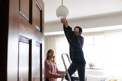 Young woman adjusting pendant light with friend at home