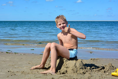 Little blond boy with swimming trunks sits laughing on a pile of sand on the beach,  with thumbs up