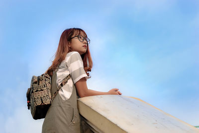 Low angle view of thoughtful woman with backpack standing against blue sky