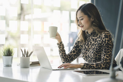 Businesswoman holding coffee cup while using laptop on desk in office