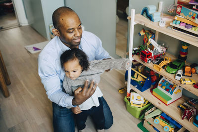 High angle view of father dressing daughter while kneeling by toys on shelf at home