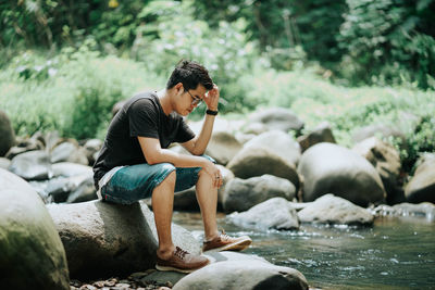 Side view of man sitting on rock by river