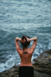 Rear view of shirtless young woman standing on rock at beach