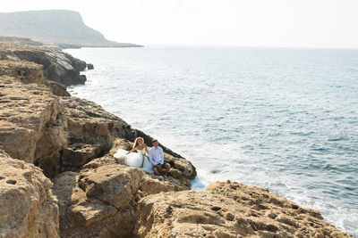 People sitting on rock by sea against sky