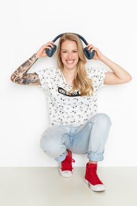 Portrait of smiling woman holding headphones while crouching against wall