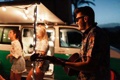 Man playing guitar for friends by illuminated campervan at night