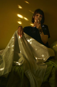 Mid adult woman smoking cigarette while sitting on bed against wall at home