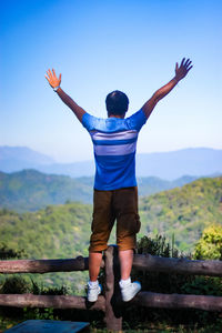 Rear view of man with arms raised standing on railing against mountains