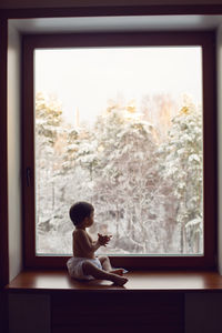 Baby boy in bath towel after washing sitting on the windowsill at the big window in winter