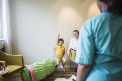 Rear view of doctor while boy with hand raised sitting in hospital