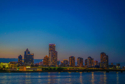 Illuminated buildings by river against blue sky