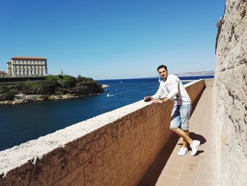 Full length portrait of young man standing by sea against clear blue sky