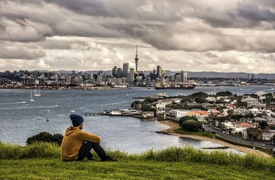 Rear view of man sitting on grass while looking at town against cloudy sky