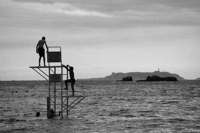 Men climbing diving platform amidst sea against sky