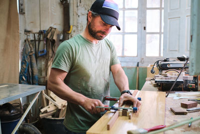 Attentive bearded male woodworker in cap gluing and clamping wooden blocks while working against window in workshop