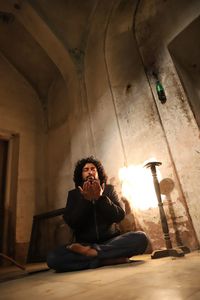 Low angle view of young man praying while sitting on floor in room