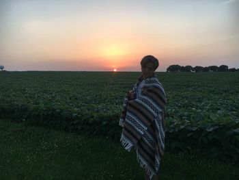 Man wrapped in blanket while standing on field against sky during sunset