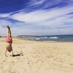 Boy doing handstand on sand at beach against sky during sunny day
