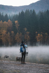 Rear view of man standing by lake in forest during foggy weather