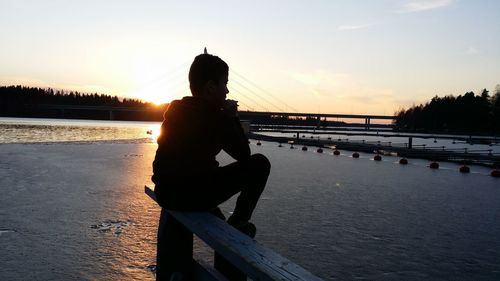 Rear view of man sitting on beach against sky during sunset