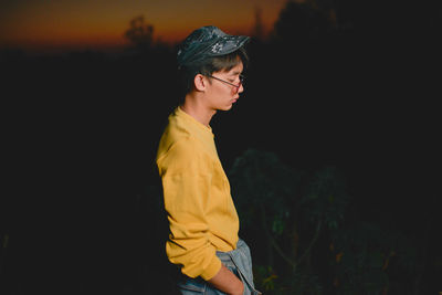 Side view of young man wearing sunglasses standing against sky at dusk